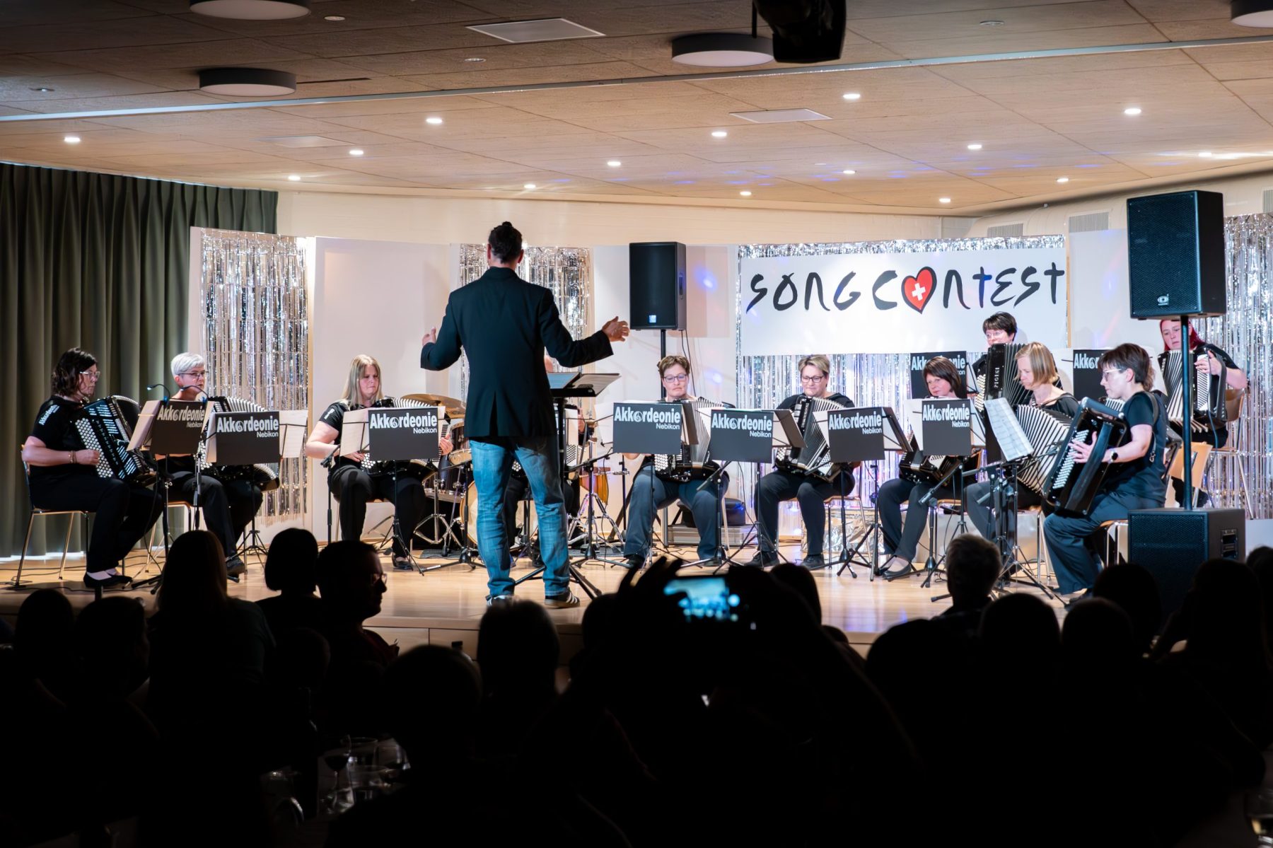 Die Akkordeonie Nebikon auf der Song-Contest-Bühne: das Ensemble in Schwarz, der Dirigent davor, Lametta-Wand und Publikum im Dunkeln.